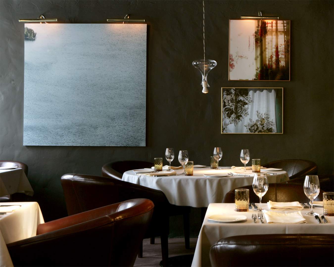 Tables set with tablecloths, wine glasses, and cutlery in front of a grey wall with art hanging on it at Le Club Chasse et Peche, one of the best special occasions restaurants in Montreal