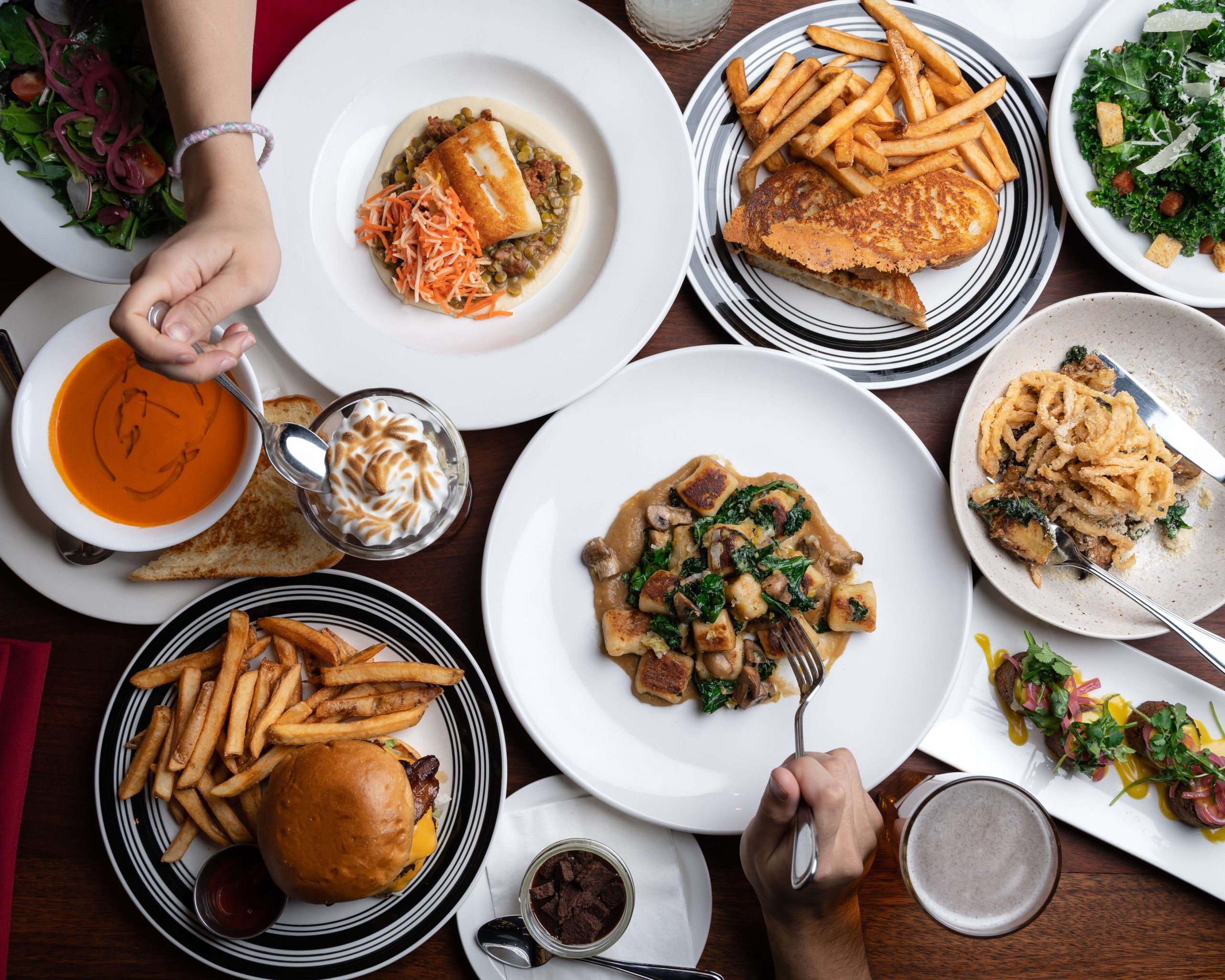 A table filled with an array of dishes, from burgers with fries to pasta, sandwiches, soups, small bites and desserts