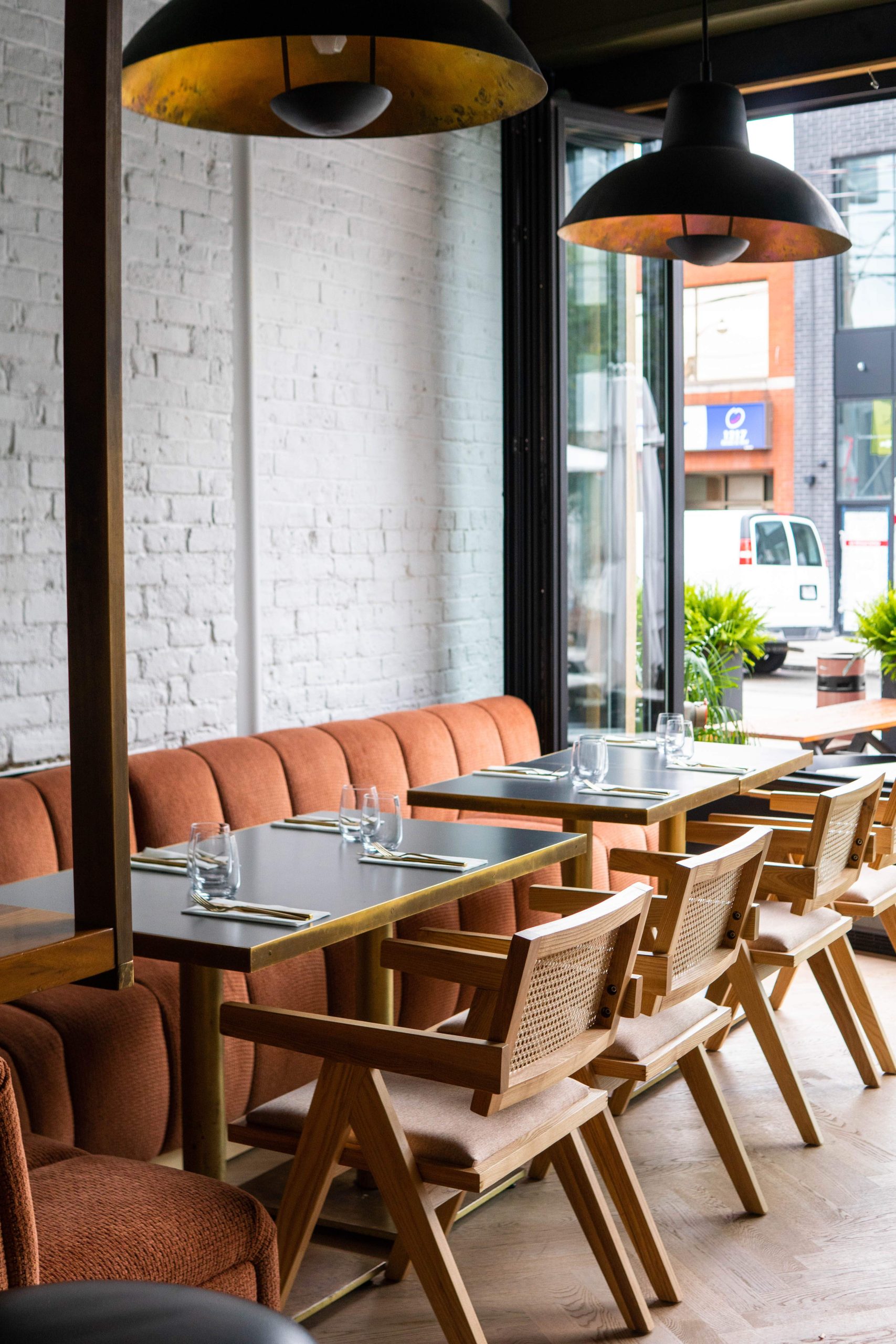 A restaurant with white brick walls, pink banquettes, dining tables, wooden chairs and a glass window