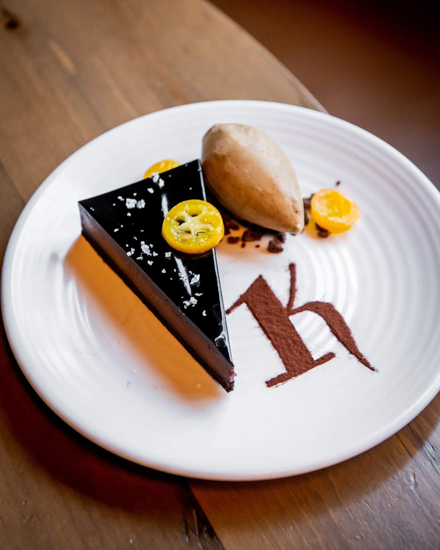 A triangle-shaped chocolate bar dessert with a scoop of ice cream and the design of the letter K made with cacao powder, on a white plate