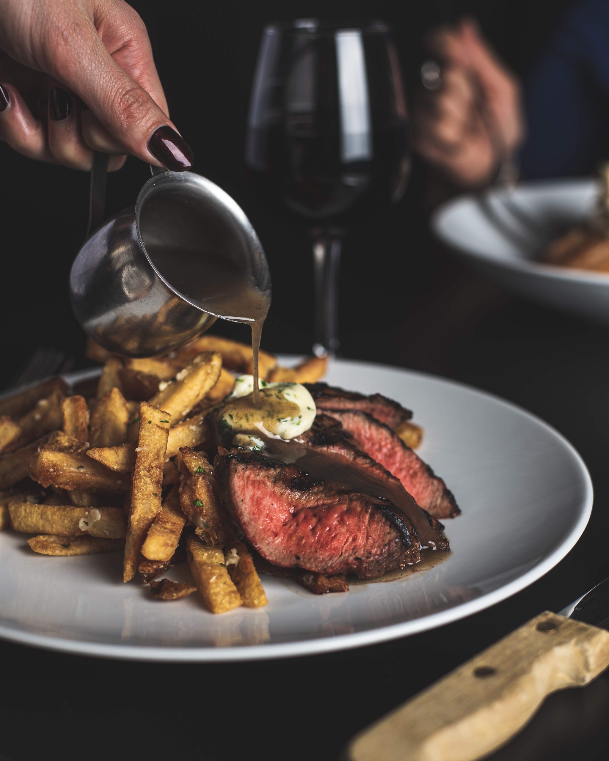 A person pouring sauce on a sliced steak with fries on a white plate.