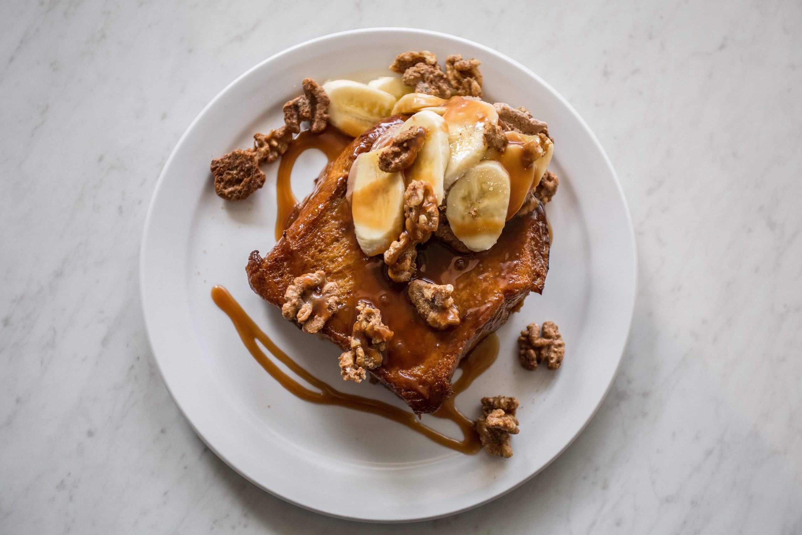 A French toast dish with nuts, banana slices and syrup on a white plate