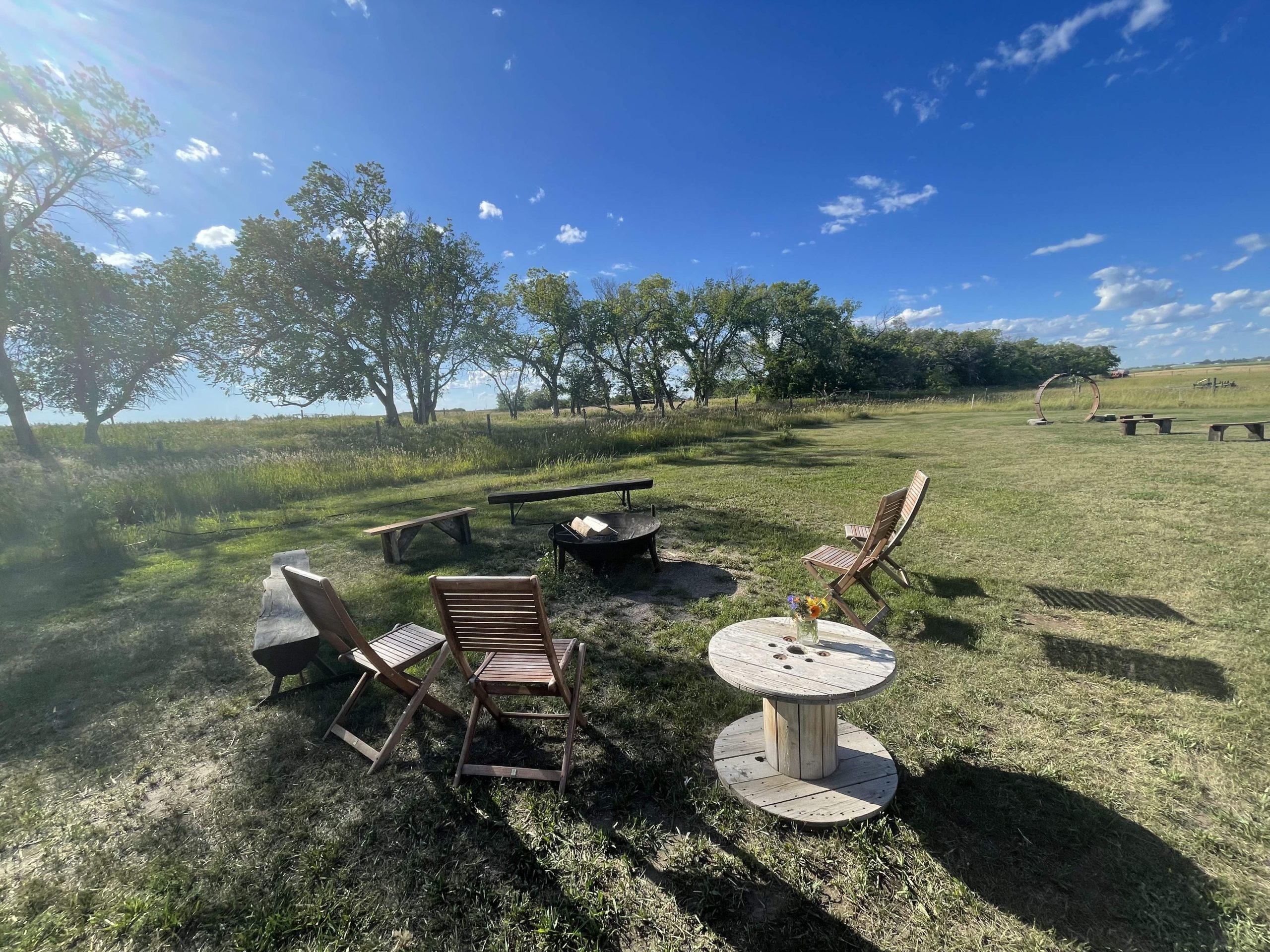 A landscape of the countryside with a wooden outdoor table and four chairs, trees and a blue sky