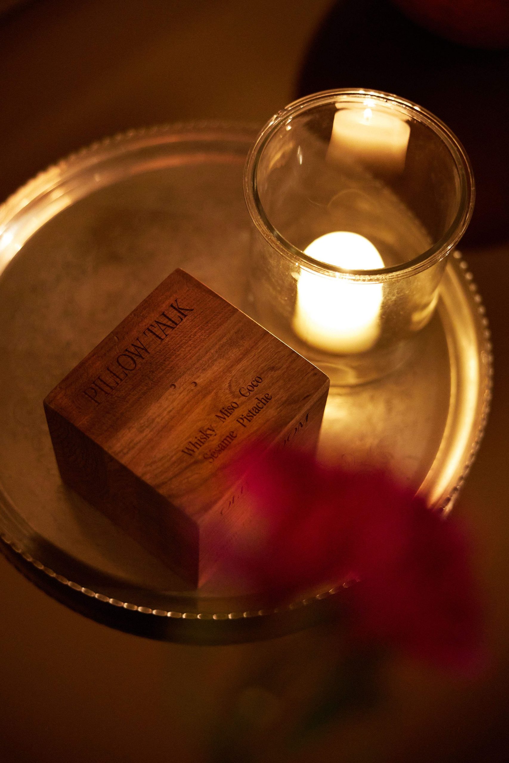 A wooden box on a silver tray next to a candle, that says 'Pillowtalk' and 'Whisky Miso Coco Sésame Pistache'