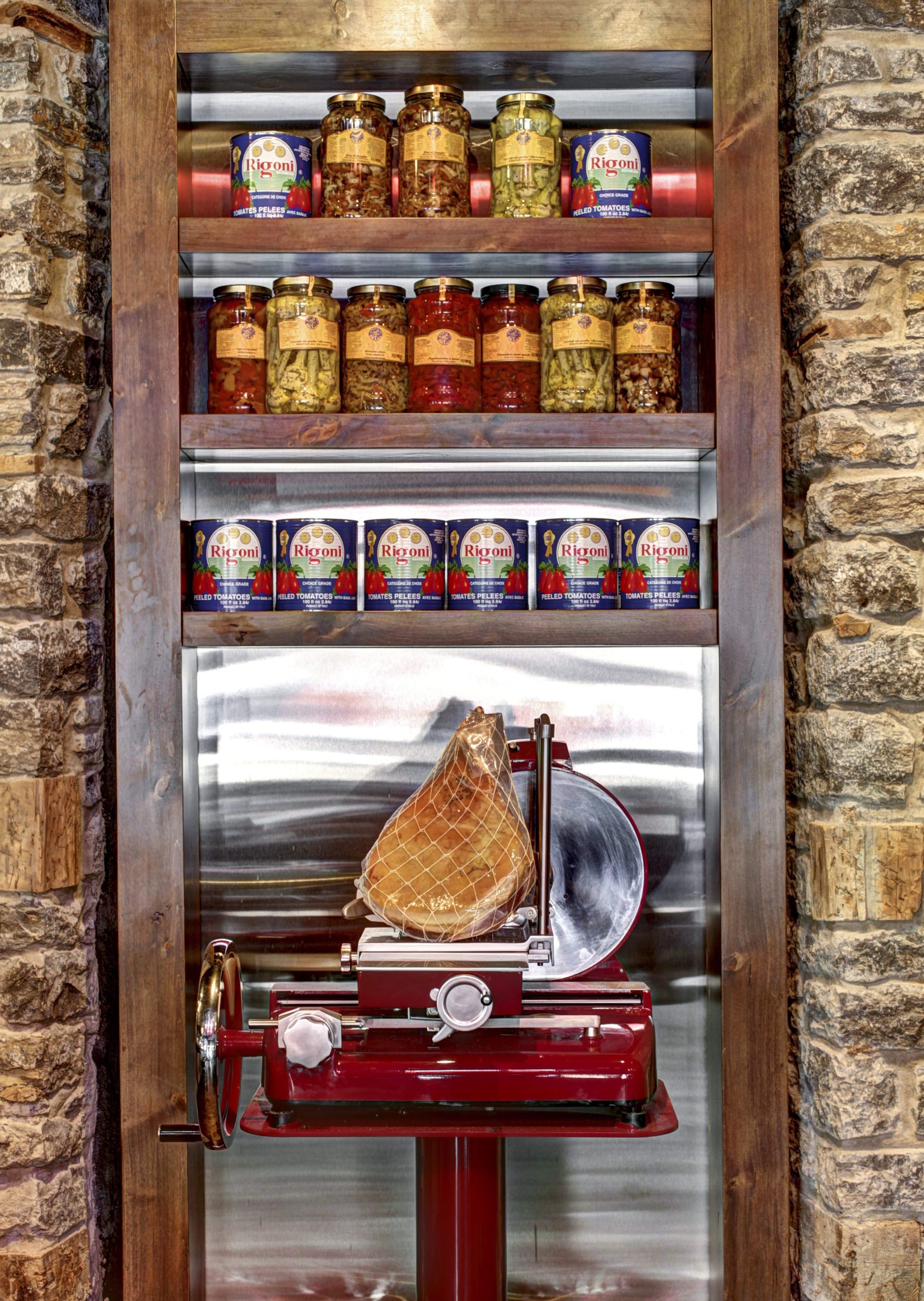 Des étagères remplies de conserves de tomates italiennes, d'artichauts et d'antipasti mixtes avec un jambon sur une trancheuse rouge.