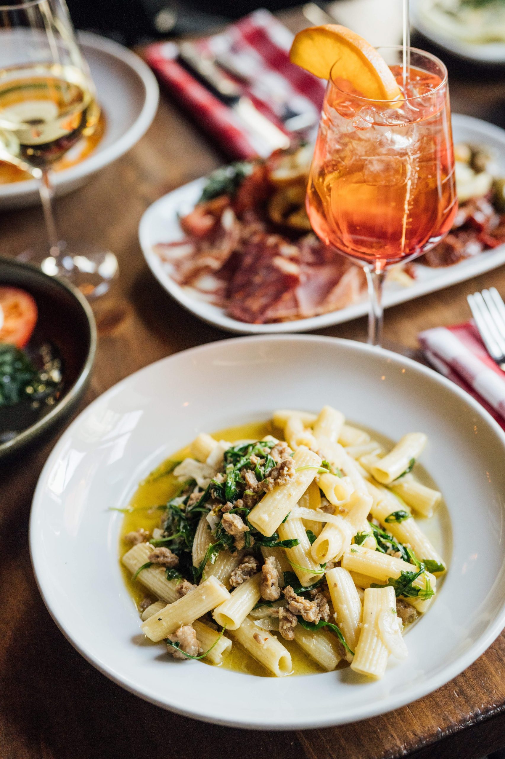 Un plat de pâtes rigatoni avec saucisses et roquette sur une assiette blanche avec un spritz et de petites assiettes en arrière-plan.