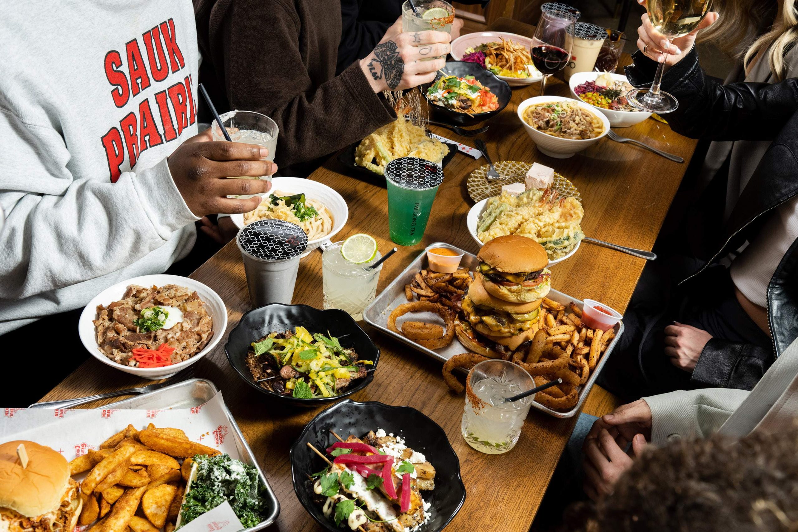 Diners feast on an array of dishes around a restaurant table, featuring burgers, salads, fries, onion rings, bubble teas and cocktails, at Chefs Hall, a food market in Toronto