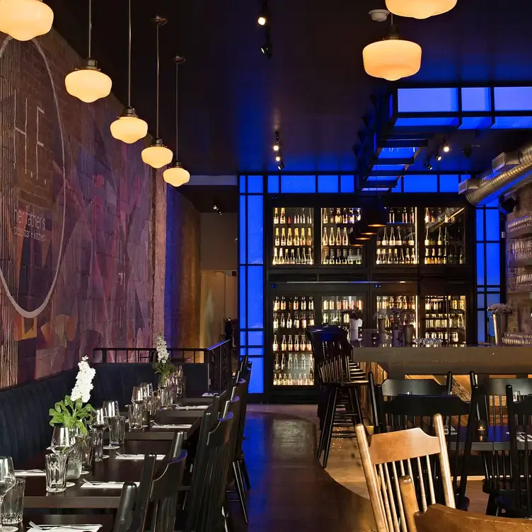 Dimly-lit interiors with hanging lights, tables and chairs lined up and a wine cooler at Her Father's Cider Bar + Kitchen