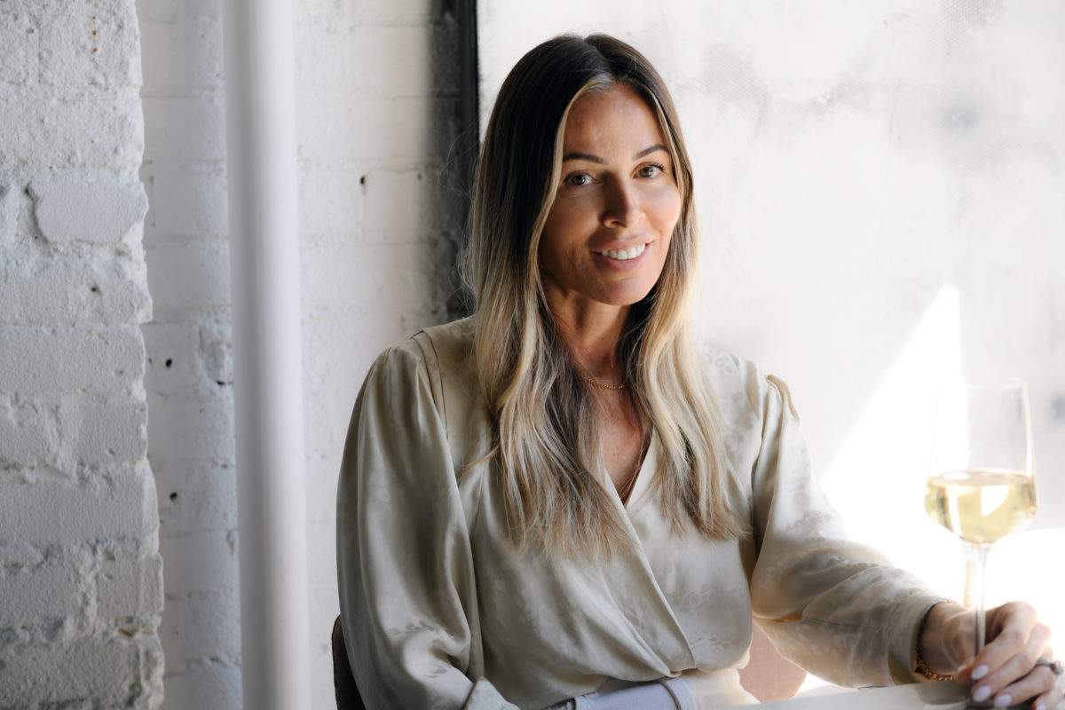 Toronto restaurateur Jenny Coburn sitting in a beige blouse holding a glass of white wine.