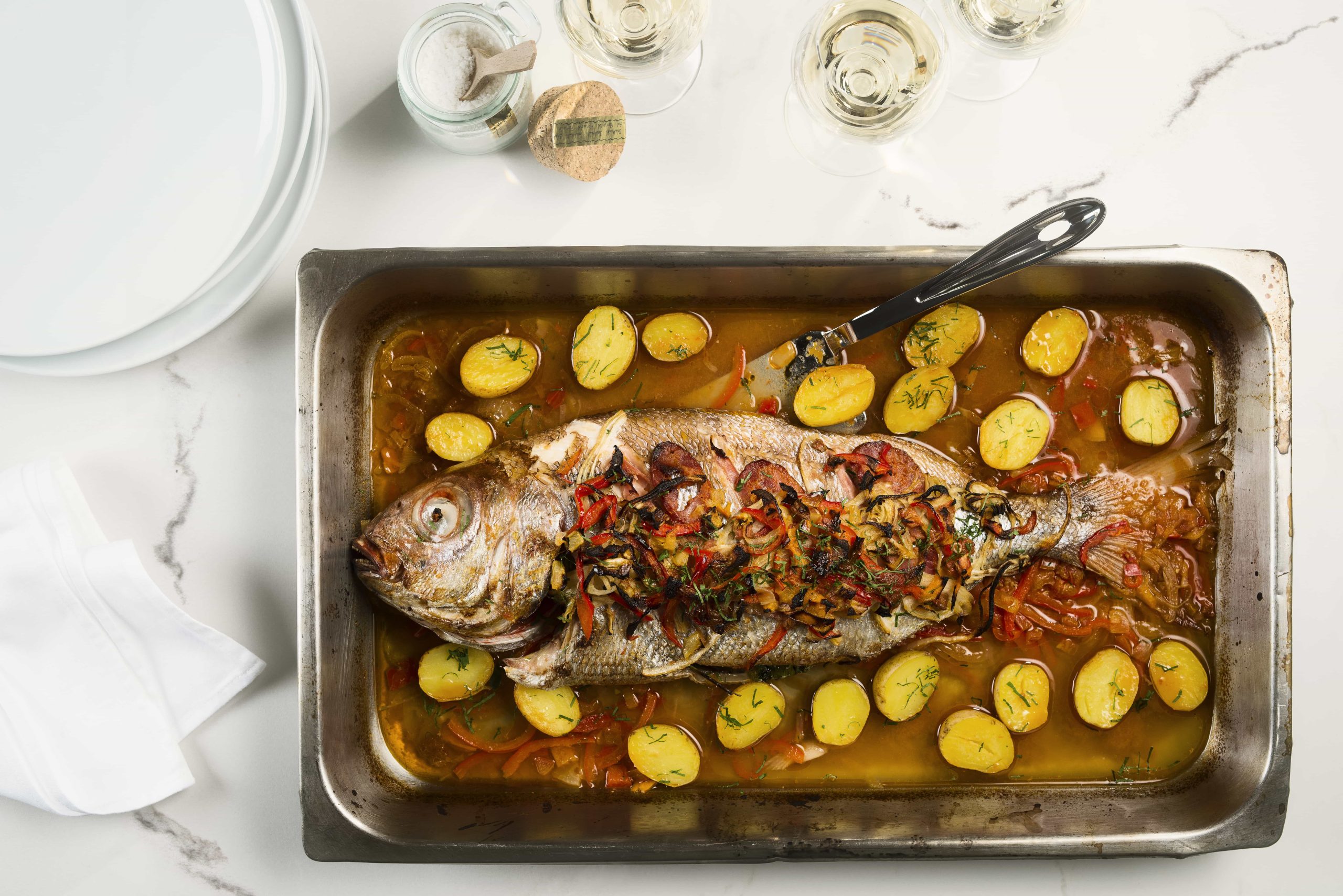 A metal tray with a whole fish surrounded by sliced potatoes at Montreal restaurant Ferreira Café.