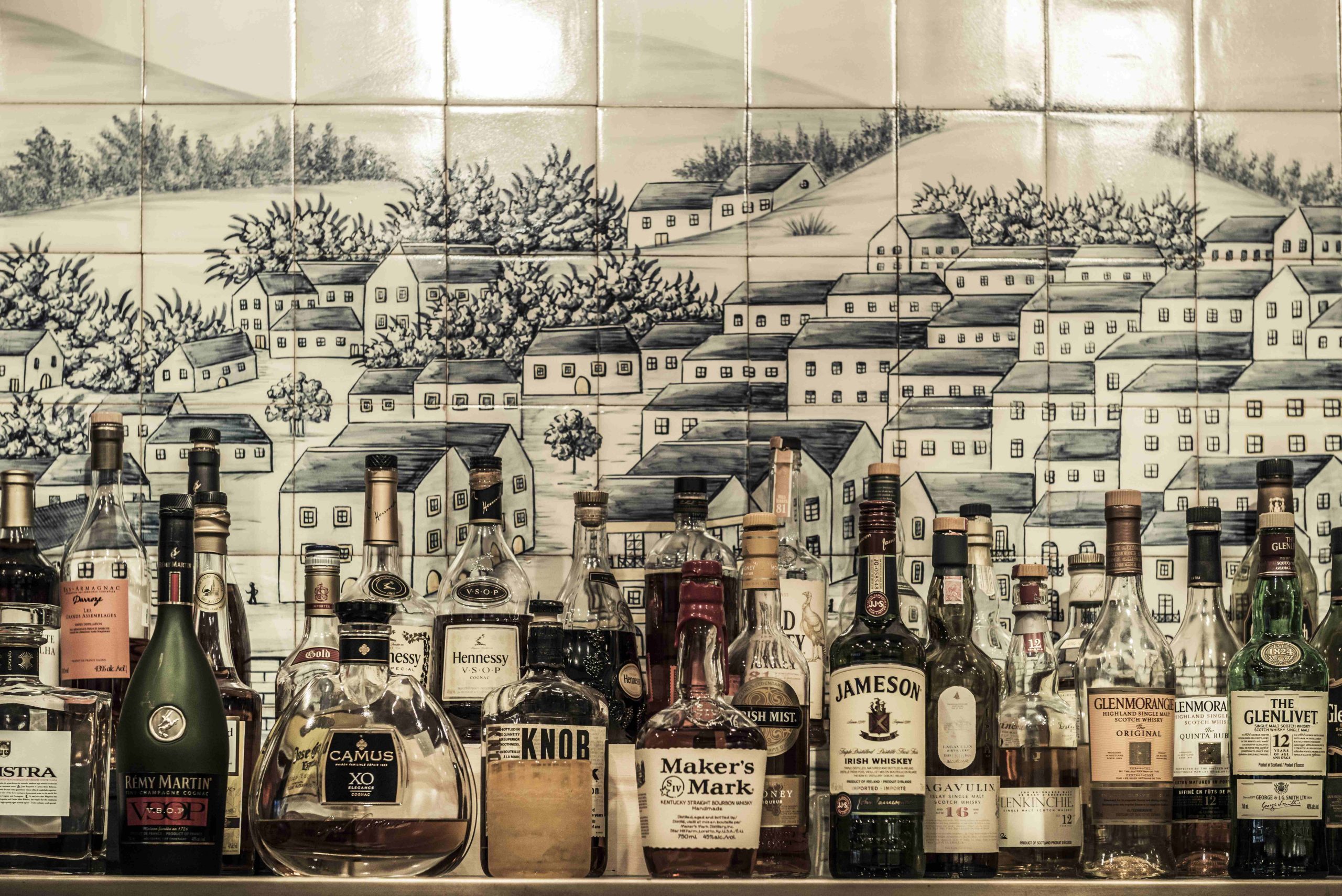 Un assortiment de bouteilles d'alcool sur une tablette devant un mur carrelé avec des images de maisons peintes sur les carreaux au restaurant montréalais Ferreira Café.