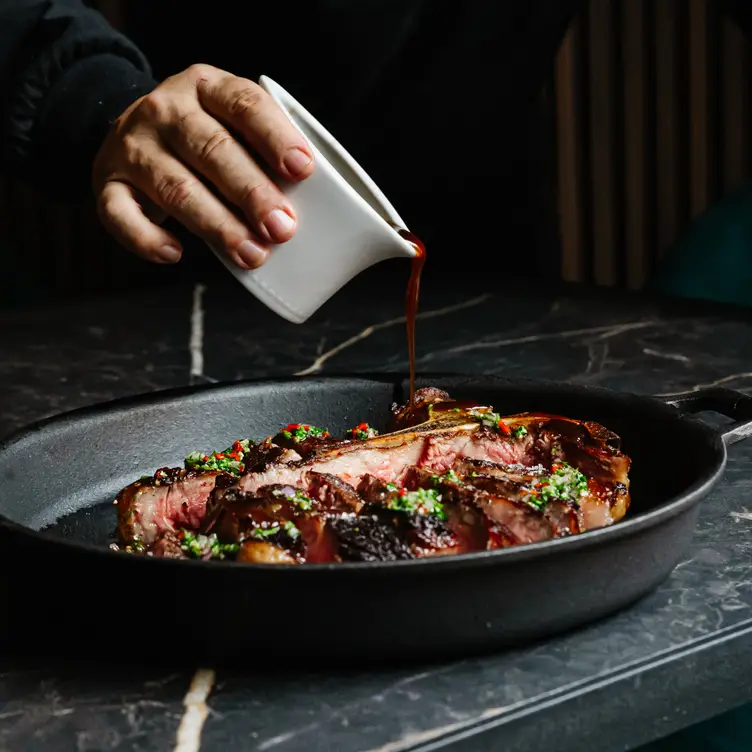 Une main verset une sauce brune sur un steak à la florentine découpé en tranches dans un plat en fonte chez L’Osso