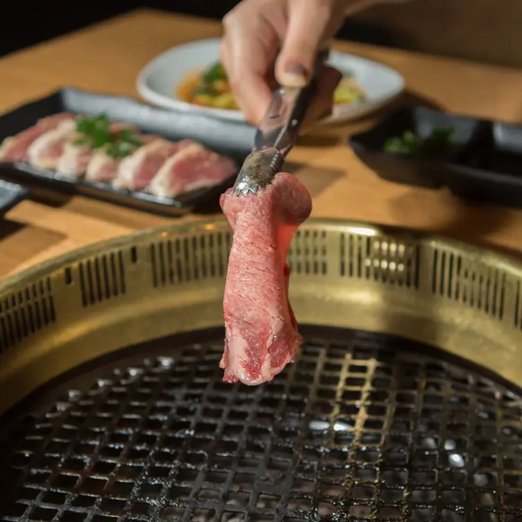 Du carpaccio de bœuf tenu par une pince à barbecue au-dessus du grill et une assiette de tranches de porc de chez Gyu-Kaku.