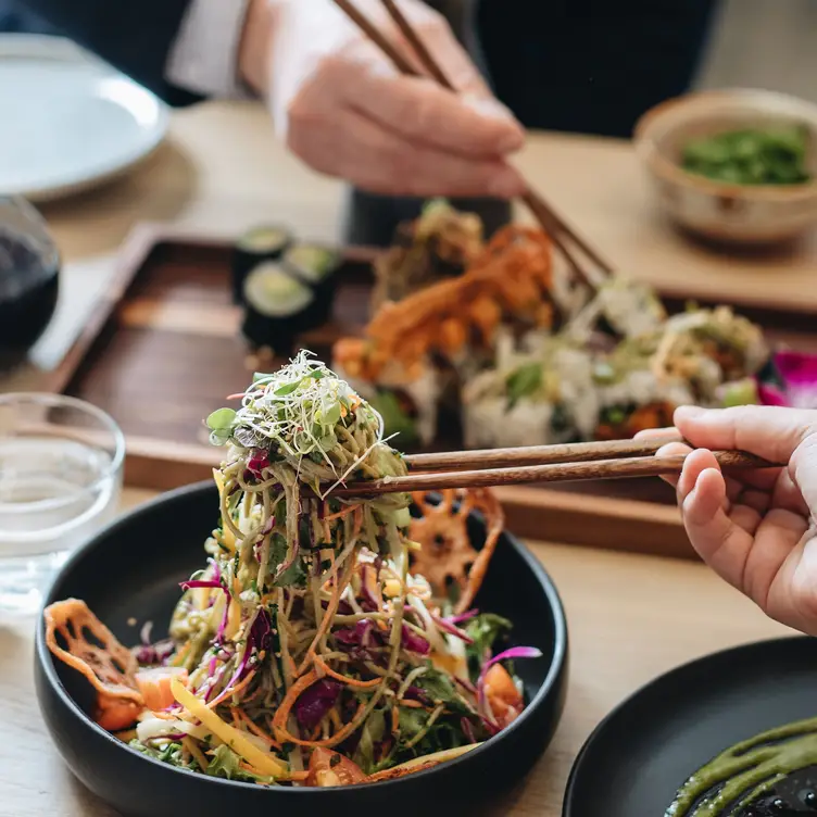 Une main tenant des nouilles d’une salade aux légumes avec des baguettes devant un plateau de makis de chez Bloom Sushi, l’un des meilleurs restaurants japonais de Montréal.