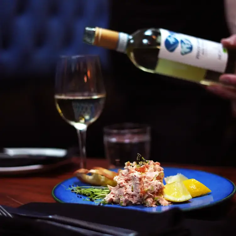 Une assiette de tartare de saumon devant une main servant un verre de vin blanc chez Le Polisson, l’un des meilleurs restaurants français à Montréal.