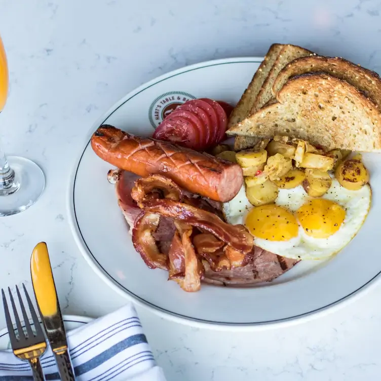 A plate with sausages, fried eggs, bacon, grilled ham, potatoes, tomatoes, and accompanied by toast at Hart’s Table & Bar, one of the best family restaurants in Edmonton.