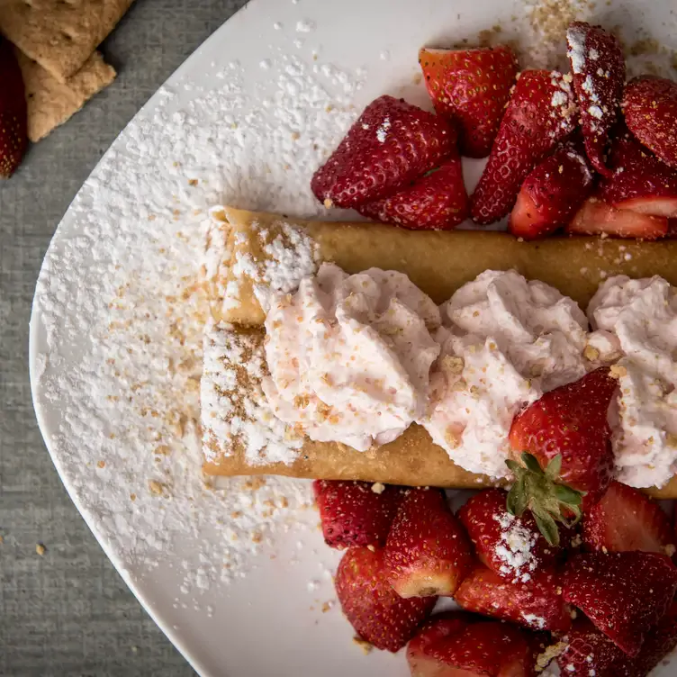 Une crêpe roulée dans une assiette avec du sucre glace, des fraises et de la crème fouettée rosée sur le dessus de chez Crêperie Chez Suzette Inc.