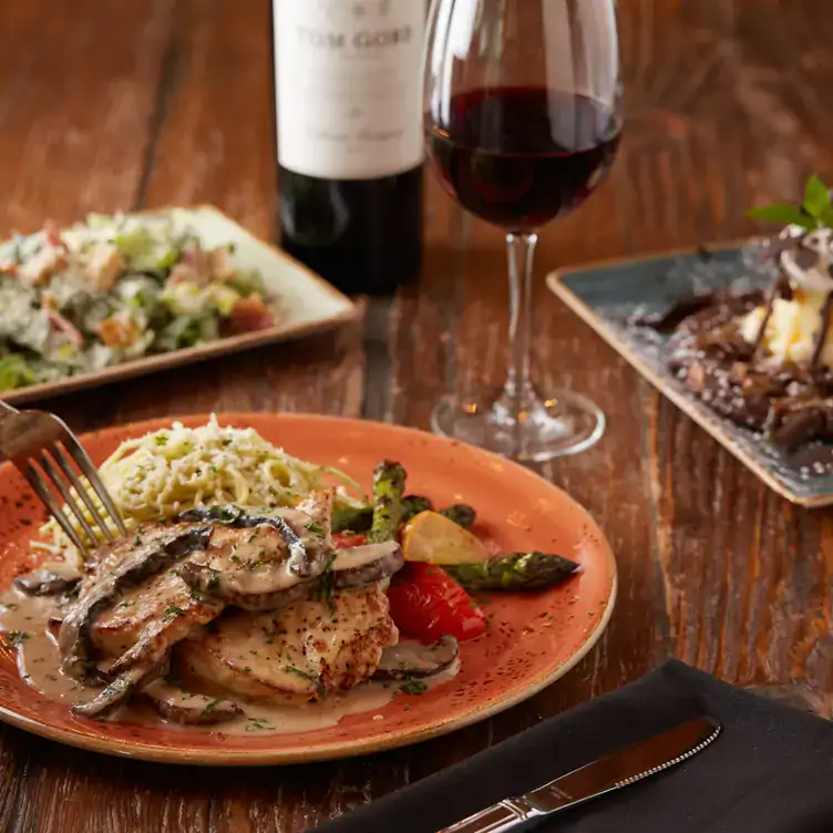 A chicken escalope on a plate with pasta, mushrooms, and other vegetables, in front of a glass and a bottle of red wine from Milestones Grill + Bar