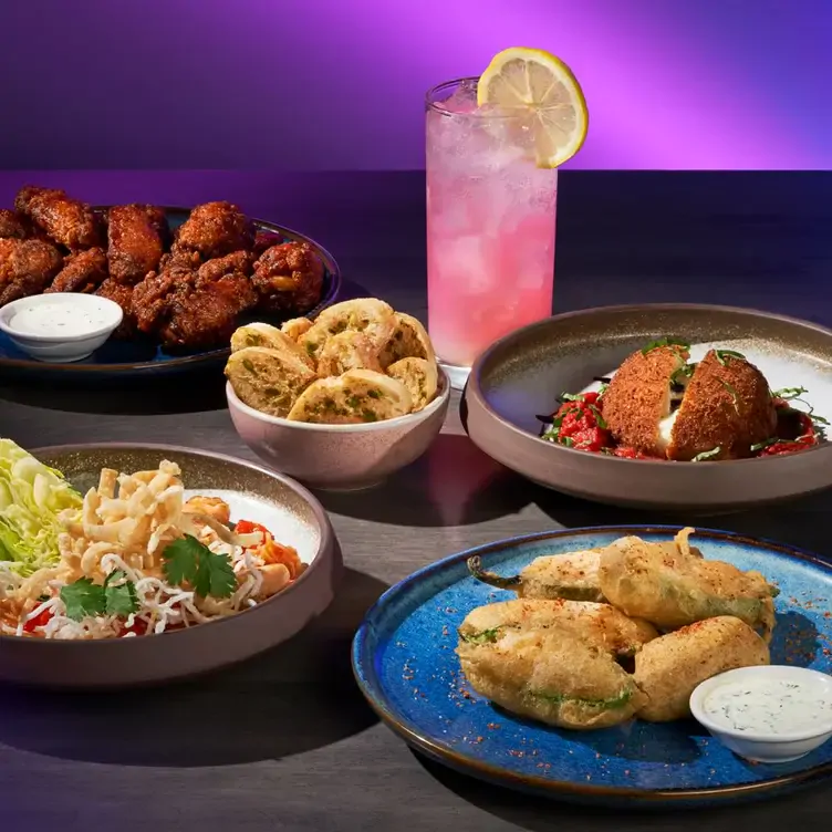 Four different plates with chicken wings, a salad, fried food, and a cheese crisp, with a bowl of seasoned bread in the center and a glass of pink drink from The Rec Room, one of the best family restaurants in Edmonton.