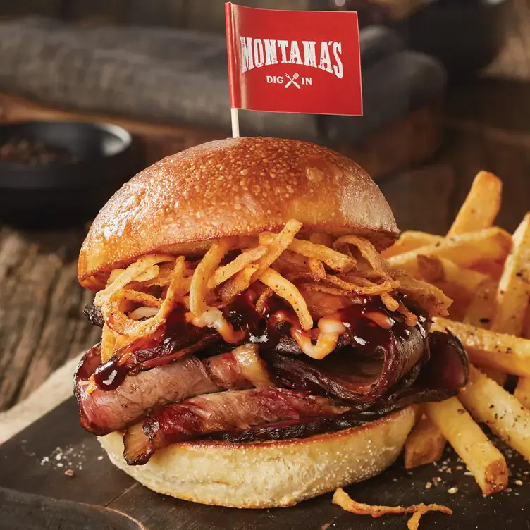 A burger made with fried onions, smoked meat, and bacon in a bun, with a small Montana's flag planted in it from Montana's BBQ & Bar, one of the best family restaurants in Edmonton.