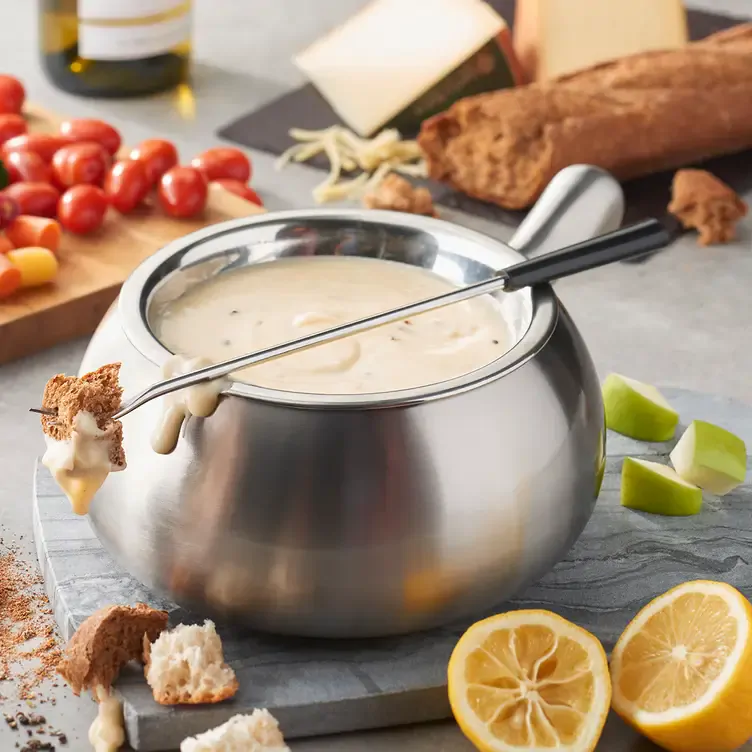 A pot of creamy fondue with a fondue skewer holding a piece of bread, all on a table filled with fruits and vegetables at The Melting Pot, one of the best family restaurants in Edmonton.