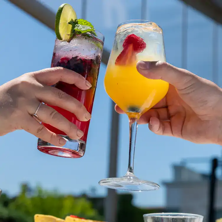 Deux mains tenant des verres de cocktail de différentes couleurs sous un ciel ensoleillé chez Perché, l’un des meilleurs restaurants sur le toit à Montréal.