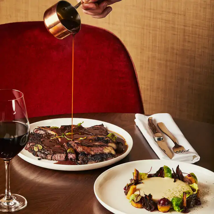 Sauce being poured over a steak on a table with wine and a dessert at Café Holt Ogilvy, one of the best fancy restaurants in Montreal.