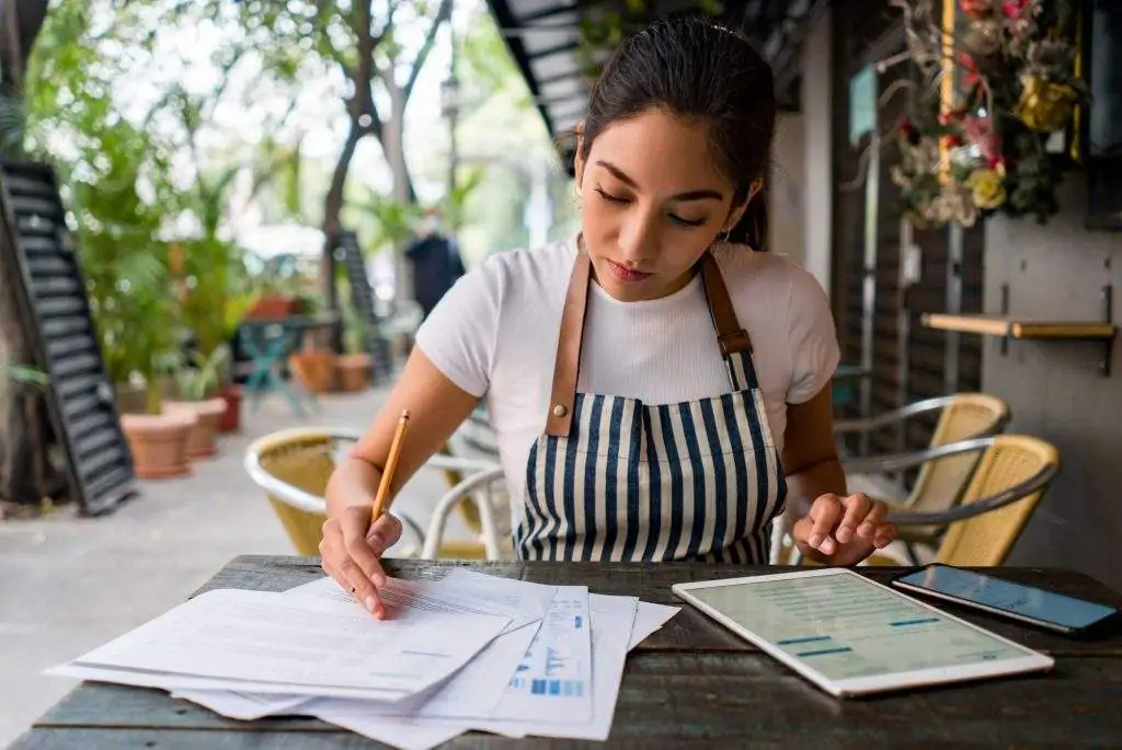 A waitress sitting in the outdoor dining area of a restaurant, wearing an apron and writing on paper while using a tablet